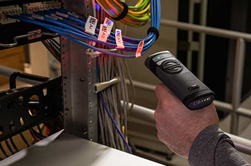 A person uses a Brady V4500 handheld barcode scanner to scan a label on a metal shelf. The scanner emits a red light onto the label, which contains a barcode.