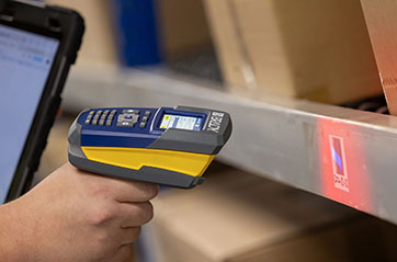 A person uses a Brady V4500 handheld barcode scanner to scan a label on a metal shelf. The scanner emits a red light onto the label, which contains a barcode.