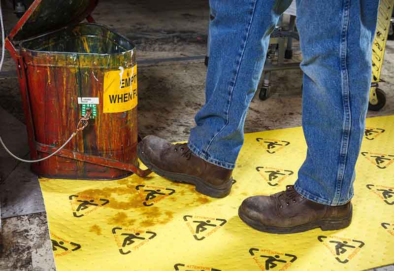 A worker in blue jeans applies a yellow spill kit safety mat to clean up a chemical hazard on a construction site.